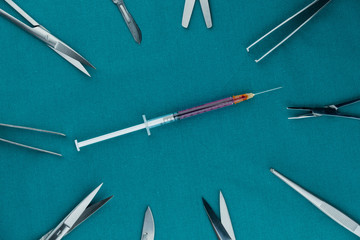 Top view of surgical instrument, liquid parenteral products in syringe with scalpel, tweezers, scissors on surgical green drape fabric.