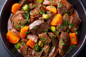 Beef meat and vegetables stew in black bowl. Dark background. Close up.