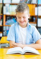 Preparing to exams. Cheerful boy reading a book and smiling while sitting on the chear at the library