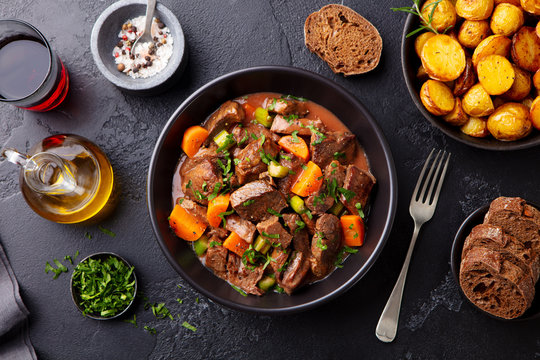 Beef meat and vegetables stew in black bowl with roasted baby potatoes. Dark background. Top view.