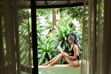 Young beautiful woman with towel on head sitting on the edge of bath and reading book after taking shower. Bali. Thailand. Koh Chang. Summer vacation.