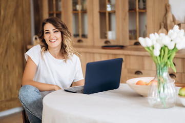 Naklejka premium Freelancer. Young positive girl working at home in a laptop while sitting at a table in the kitchen