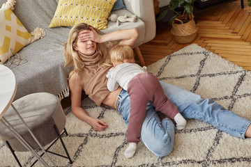 Blonde Caucasian woman in jeans is sitting on carpet her little baby child in a bright room with windows in Scandinavian style breast feeding exhausted - 328044672