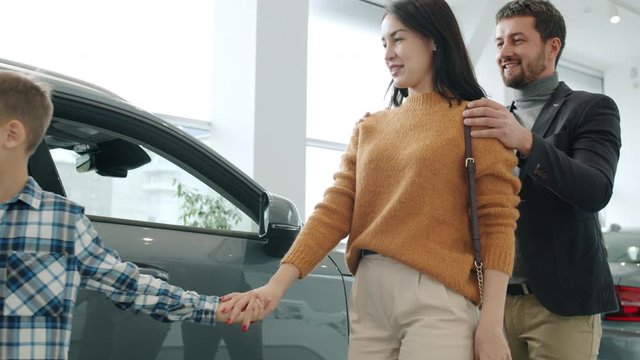 Young family mother, father and child are buying car making surprise for woman hugging enjoying purchase. Excitement, people and modern lifestyle concept.