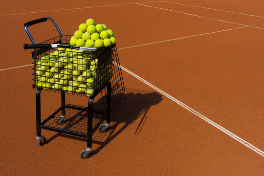 Blaсk basket-cart with tennis balls and a rocket with a pyramid of balls on a tennis clay court. Sport, recreation and training concept. Background for tennis school banner. Red court and yellow balls