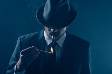 Mafioso with covered eyes with felt hat lighting cigar on dark blue background - obrazy, fototapety, plakaty
