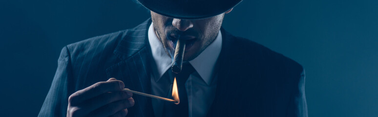 Mafioso lighting cigar with match on dark blue, panoramic shot