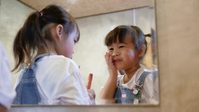 Cute sibling child girl doing makeup and having fun applying lipstick together at a mirror in room.