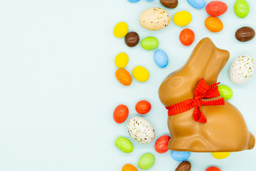 Happy easter card. Easter milk chocolate bunny, rabbit with red bow on a neck. There are colourful candies and small eggs around. Blue background. Isolated. Copy space on the left side.