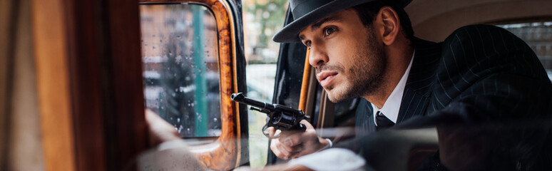 Selective focus of gangster sitting in ambush with gun in retro car, panoramic shot