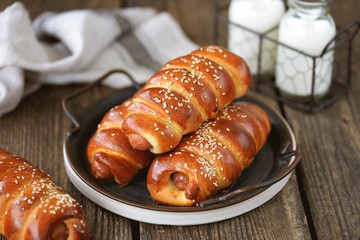 Homemade sausages in yeast dough with sesame seeds and milk on a wooden background.