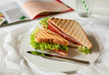 Sandwich with crispy toasts, slices of pork ham, cheese, tomato, salad leaf on the white plate, glass of water - tasty healthy time break concept.