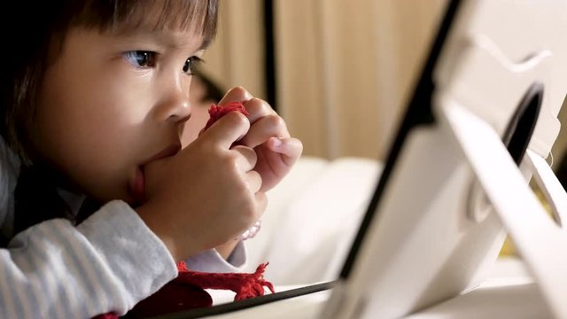 Adorable little girl lying on bed with sucking her thumb and looking on a digital tablet in  bedroom. 