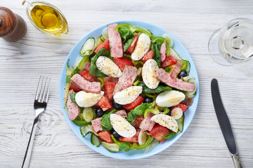 A classic Nicoise salad is on a blue plate. Nearby a knife, fork, a glass of white wine, pepper, sauce. View from above. Close-up.