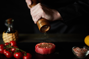 Chef sprinkles pepper with tuna steak. Hands of the cook on a dark background. Close-up