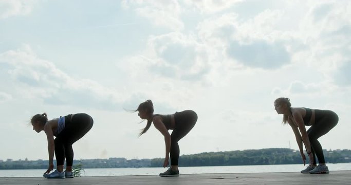 Three athletic young women working out together on a wooden deck overlooking water leaping, stretching and squatting in a side view against a high key sky in a health and fitness concept