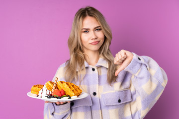 Teenager Russian girl holding waffles isolated on purple background showing thumb down