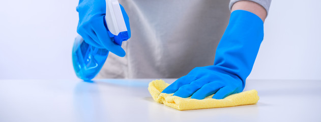 Young woman housekeeper is doing cleaning white table in apron with blue gloves, spray cleaner, wet...