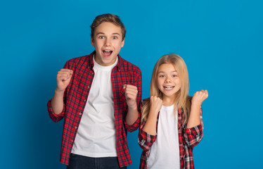 Excited boy and girl celebrating success, raising fists on blue background