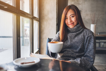 Closeup image of a beautiful asian woman showing and drinking coffee while reading a book