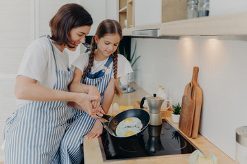 Busy mother and daughter wear striped aprons, pose at kitchen near cooker, fry eggs on pan, prepare fast breakfast, enjoy domestic atmosphere. Mum teaches small kid to cook. Happy family concept