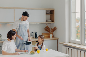 Father prepared fried eggs for family, little child holds plate and waits for breakfast. Family pose at kitchen near table, enjoy tasty meal, have glad expressions. People, eating, domestic atmosphere