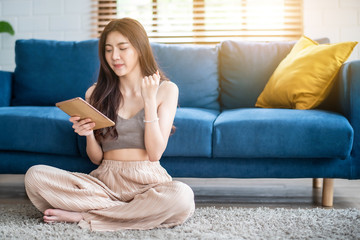 Young beautiful Asian woman sitting on blue sofa and reading web by tablet at living room in the morning