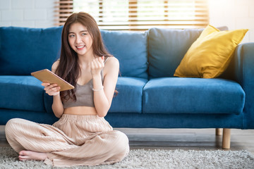 Young beautiful Asian woman sitting on blue sofa and reading web by tablet at living room in the morning