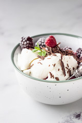 Vanilla ice cream with frozen raspberries, blackberries, chocolate and mint in a ceramic bowl, light grey background.