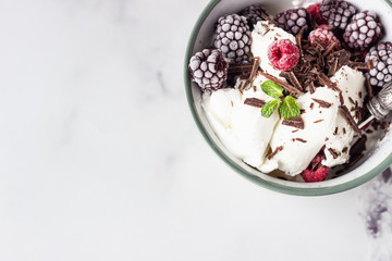 Vanilla ice cream with frozen raspberries, blackberries, chocolate and mint in a ceramic bowl, light grey background.