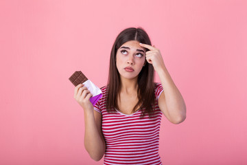 Teenage girl with acne problem holding chocolate bar against pink background. Young beautiful Woman...