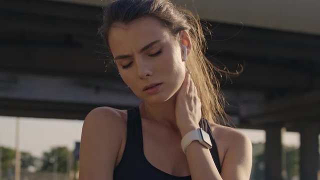A tired sporty young woman with earphones is touching her neck while having pause of training outdoors near the bridge in the city