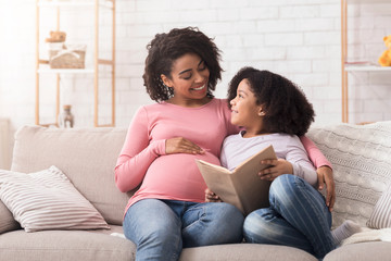 Happy African Mom And Her Daughter Reading Book In Living Room