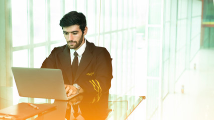 Young caucasian man with beard in airplane pilot uniform using a laptop in modern office.Handsome...
