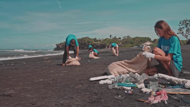 Young Woman and Friends in Blue T-shirt Collecting Trash into Eco Bags on Beach. Pollution of Seaside with Different Kinds of Garbage. Volunteers Clean Up Wastes on Ocean Coast. Ecology Protection