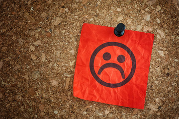 Red paper note with sad face on cork board background