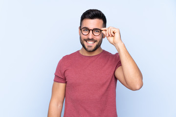 Young handsome man with beard over isolated blue background with glasses and happy