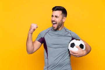 Man over isolated yellow background with soccer ball celebrating a victory