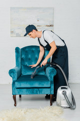 Fototapeta na wymiar side view of cleaner in uniform cleaning modern armchair with vacuum cleaner
