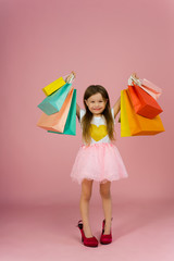 Beautiful cute little girl with long blonde hair, tulle skirt standing in mothers big shoes with colorful packages isolated on pink background. Expressing true positive emotions to camera