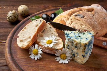 Sliced homemade ciabatta, blue gorgonzola cheese, olive branch and quail eggs. Chamomile flowers and a bread knife. Wooden rustic background.