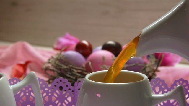 Easter table setting for the festive breakfast: a tea poured in the cup on the background of colored pink Easter eggs in the nest