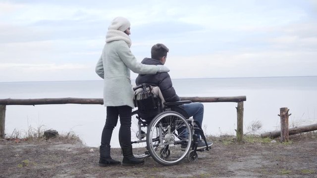 Young positive Caucasian woman stopping wheelchair with disabled boy on river bank and pointing away. Mother spending autumn day with handicapped son outdoors. Lifestyle, disability, support.