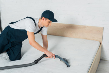 handsome cleaner in cap removing dust on mattress with vacuum cleaner