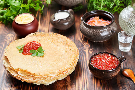 A stack of thin pancakes(blini) with red caviar. Around the pots with red fish, caviar, fresh butter and sour cream. Nearby is a decanter, and a stack of vodka. Russian holiday. Maslenitsa.
