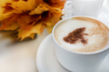 Coffee (cappuccino with cinnamon) in a white cup, is on the table. Nearby lies a bouquet of maple leaves