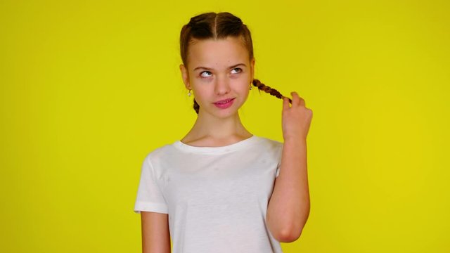 Teenage girl in a white T-shirt chews gum and twists a pigtail. Yellow background with copy space. Teenager emotions. 4k footage