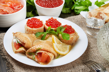 Russian thin pancakes(blini) with red fish and red caviar lie on a white plate. Near the carafe and...