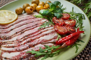 Slices of bacon with dried tomatoes, green olives, arugula, basil and lemon lie on a green plate