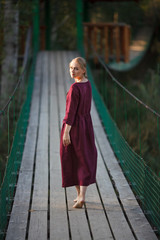 A girl in a burgundy dress walks on a suspended wooden bridge